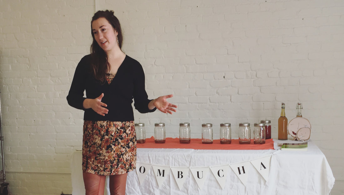 Woman teaching kombucha brewing corporate class with kombucha jars on a table in the background.