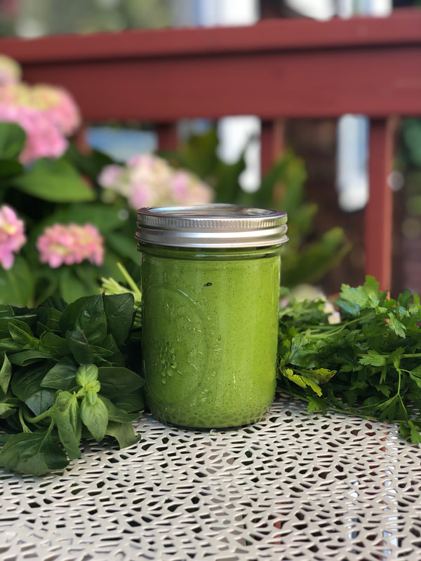 green sauce in jar on white table with basil and cilantro in background