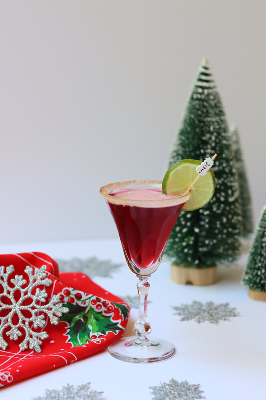 Red cranberry cosmo with orange and cinnamon sugar rim with christmas trees in the background and mistletoe napkin 
