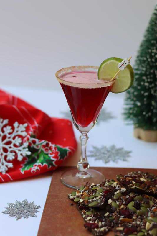 Red cranberry cosmo with sugar rim, lime slice and snowman stir stick on in winter holiday background with sweet and salty chocolate bark on cutting board