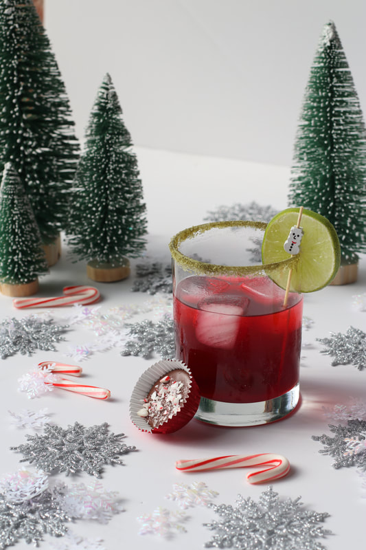 Red Margarita with matcha sugar rim, lime slice and snowman stir stick on in winter holiday background with peppermint bark bites in red foil.