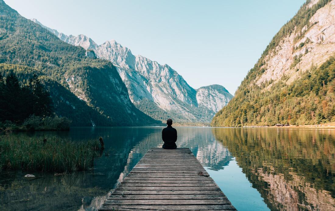 person sitting at the end of a lakeside dock overlooking beautiful mountains with green trees and white rocks